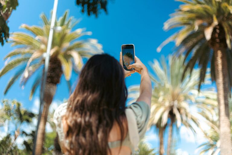 Junge Frau richtet ihr Smartphone auf Palmen vor strahlend blauem Himmel.