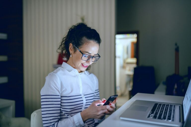 Junge Frau sitzt am Laptop in einem Zimmer und schaut auf ihr Smartphone.