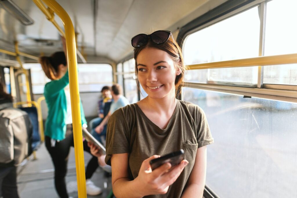 Lächelnde junge Frau mit Smartphone genießt eine Busfahrt mit Sonnenbrille im Haar